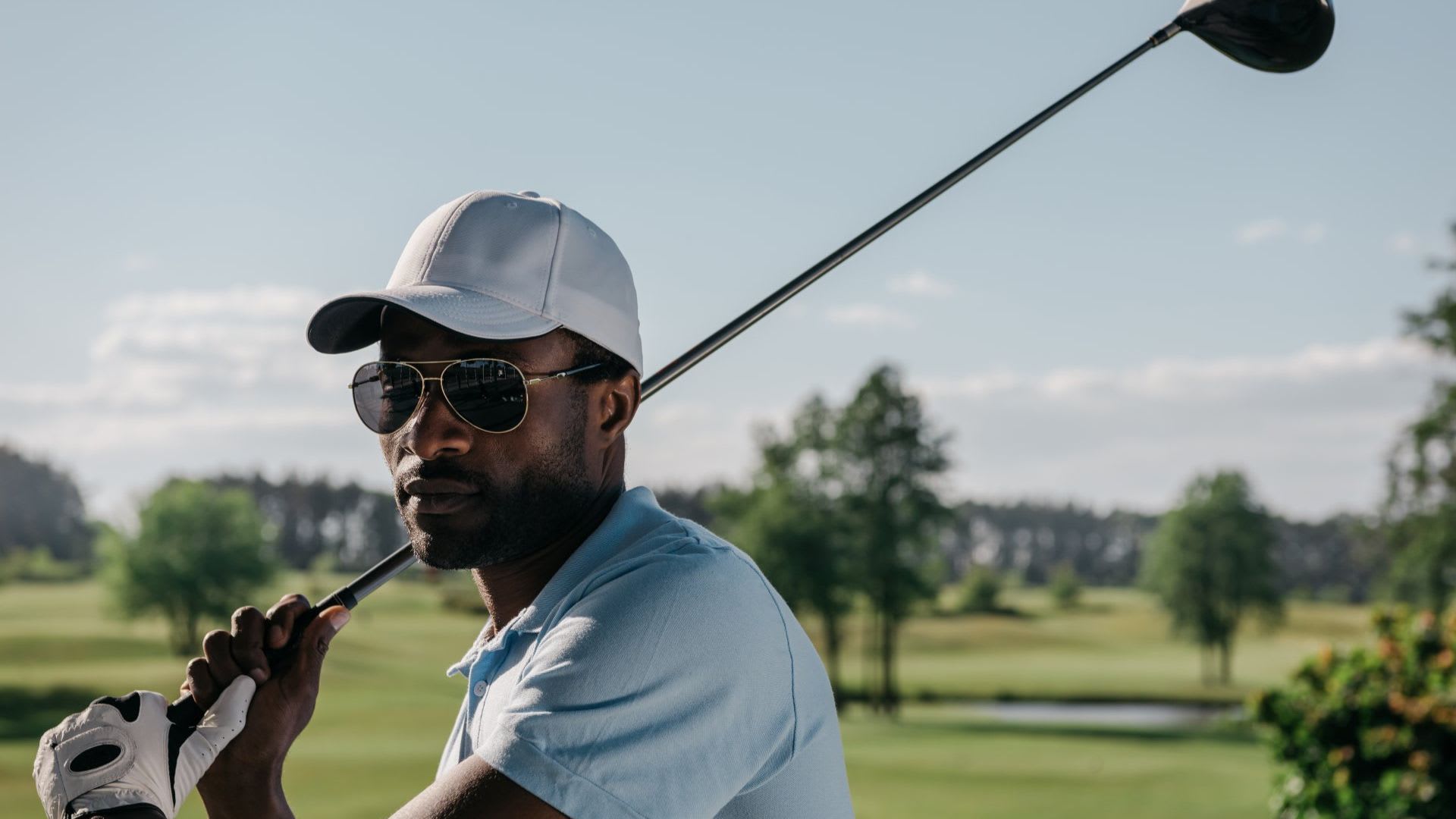 A man wearing sunglasses and a white cap holds a golf club over his shoulder on a golf course, showcasing popular golf brands, with trees and blue sky in the background.